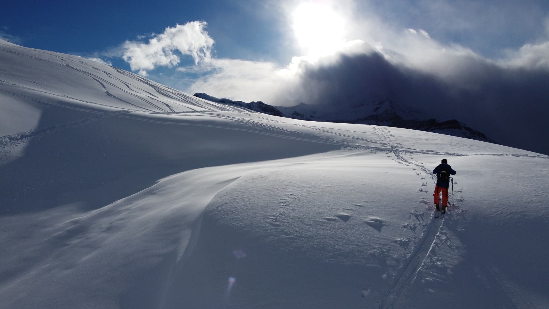 Ski tourer crossing a wide snow-covered glacier slope with the sun breaking through the clouds, Zermatt